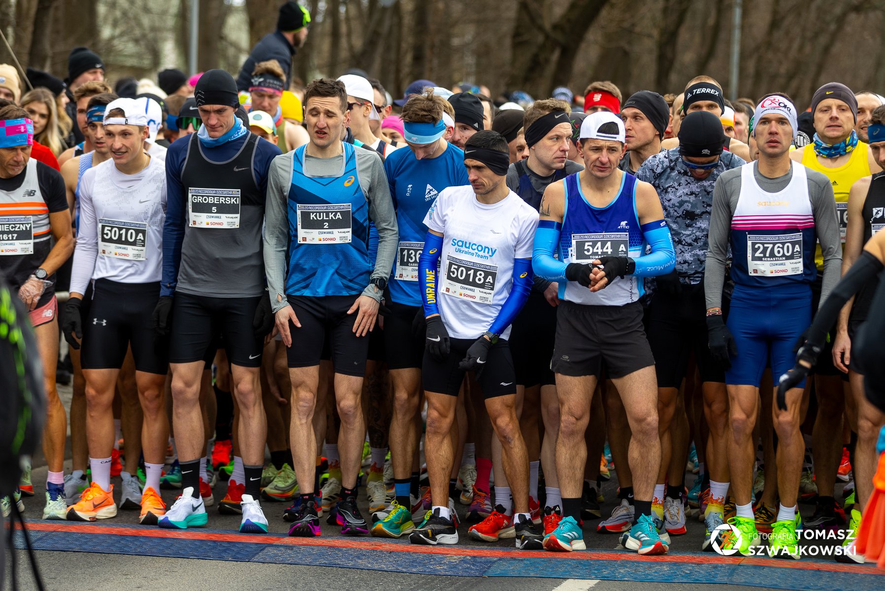 20. Recordowa Dziesiątka, fot. Tomasz Szwajkowski - grupa biegaczy przygotowuje się do startu. Wszyscy sa w krótkich spodniach, niektórzy mają czpaki na głowie i większość ma długie rękawy.