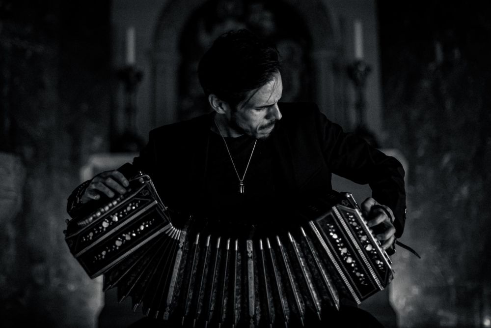 Artysta siedzi na krześle i gra na bandeonie, na twarzy ma głębokie skupienie.