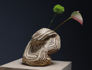 Artystyczna rzeźba ceramiczna o organicznej, warstwowej strukturze w odcieniach beżu i brązu, ustawiona na postumencie i ozdobiona dwoma pojedynczymi kwiatami.