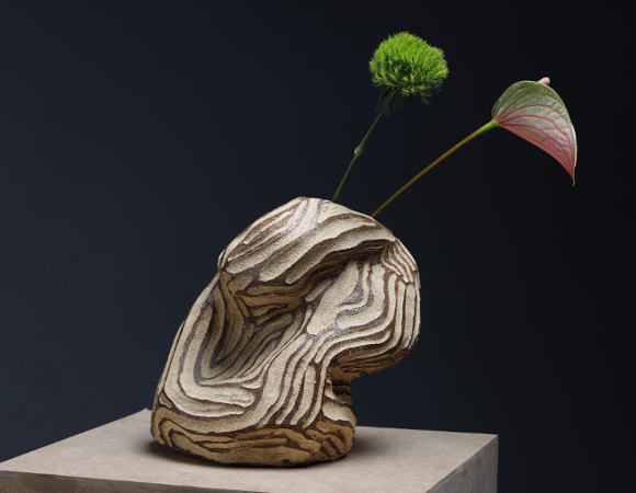 Artystyczna rzeźba ceramiczna o organicznej, warstwowej strukturze w odcieniach beżu i brązu, ustawiona na postumencie i ozdobiona dwoma pojedynczymi kwiatami.