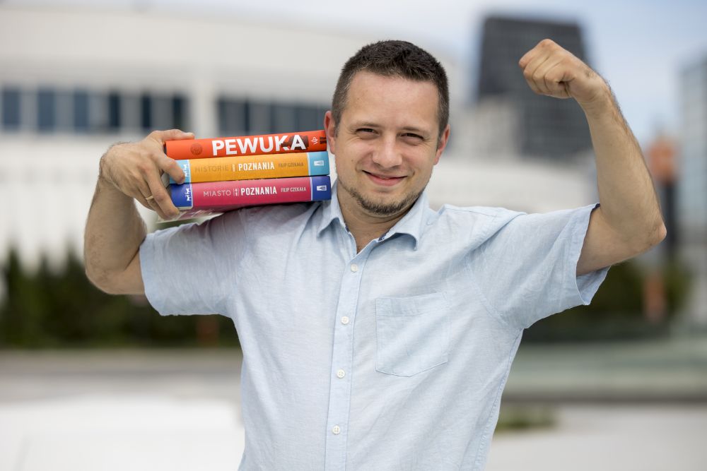 Autor siedzi po turecku i trzyma swoją książkę, pozuje na ul. Święty Marcin, za nim widać restaurację Sphinx, szyld Biedronki i nadjeżdżający tramwaj.