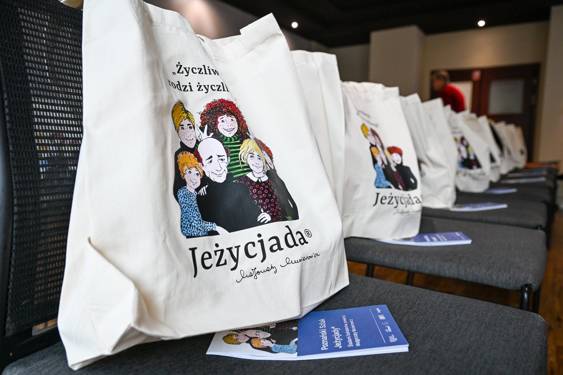 Białe bawełniane torby z rysunkową grafiką przedstawiającą rodzinę oraz napisem "Jeżycjada" i "Życzliwi rodzi życzliwi" stoją na krzesłach w sali konferencyjnej. Na krześle, na pierwszym planie, leży fioletowa ulotka.
