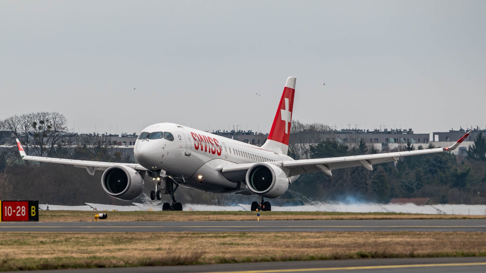 Biało-niebieski samolot pasażerski linii Lufthansa z napisem "Lufthansa" oraz logo na stateczniku pionowym kołuje na pasie startowym. W tle widoczne są zabudowania lotniskowe, maszty z oświetleniem oraz inne mniejsze samoloty zaparkowane przy terminalu. Na pierwszym planie pas startowy z oznaczeniami poziomymi.