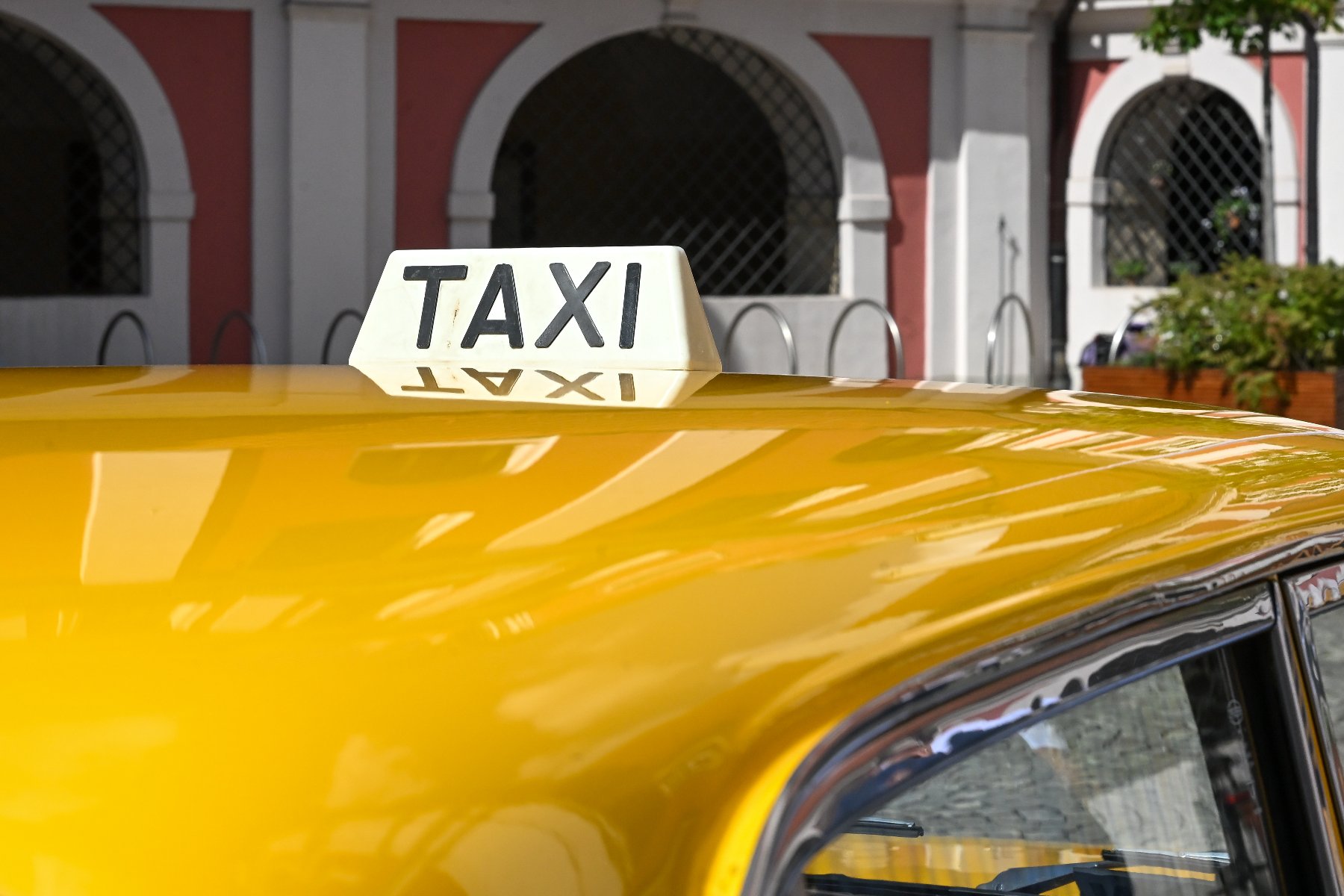 Biały kogut z czarnym napisem "TAXI" zamontowany na dachu żółtego samochodu, zaparkowanego przed zabytkowym budynkiem z arkadami.