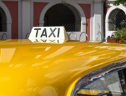 Biały kogut z czarnym napisem "TAXI" zamontowany na dachu żółtego samochodu, zaparkowanego przed zabytkowym budynkiem z arkadami.