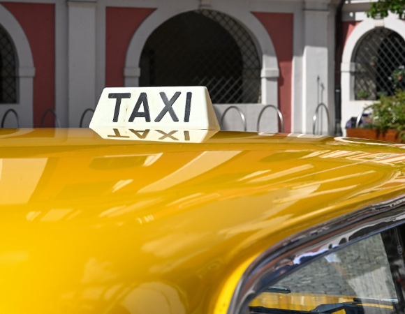 Biały kogut z czarnym napisem "TAXI" zamontowany na dachu żółtego samochodu, zaparkowanego przed zabytkowym budynkiem z arkadami.