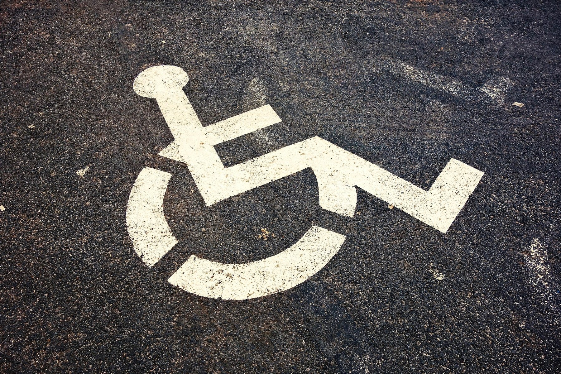 Biały piktogram wózka inwalidzkiego, symbolizujący miejsce parkingowe dla osób z niepełnosprawnościami, namalowany na ciemnym asfalcie.