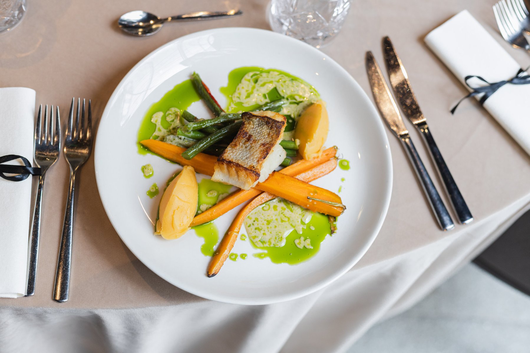 Biały talerz z daniem głównym na stole. Na talerzu kawałek smażonej ryby, marchewki, zielona fasolka i purée ziemniaczane, polane zielonym sosem. Obok talerza sztućce i serwetki.