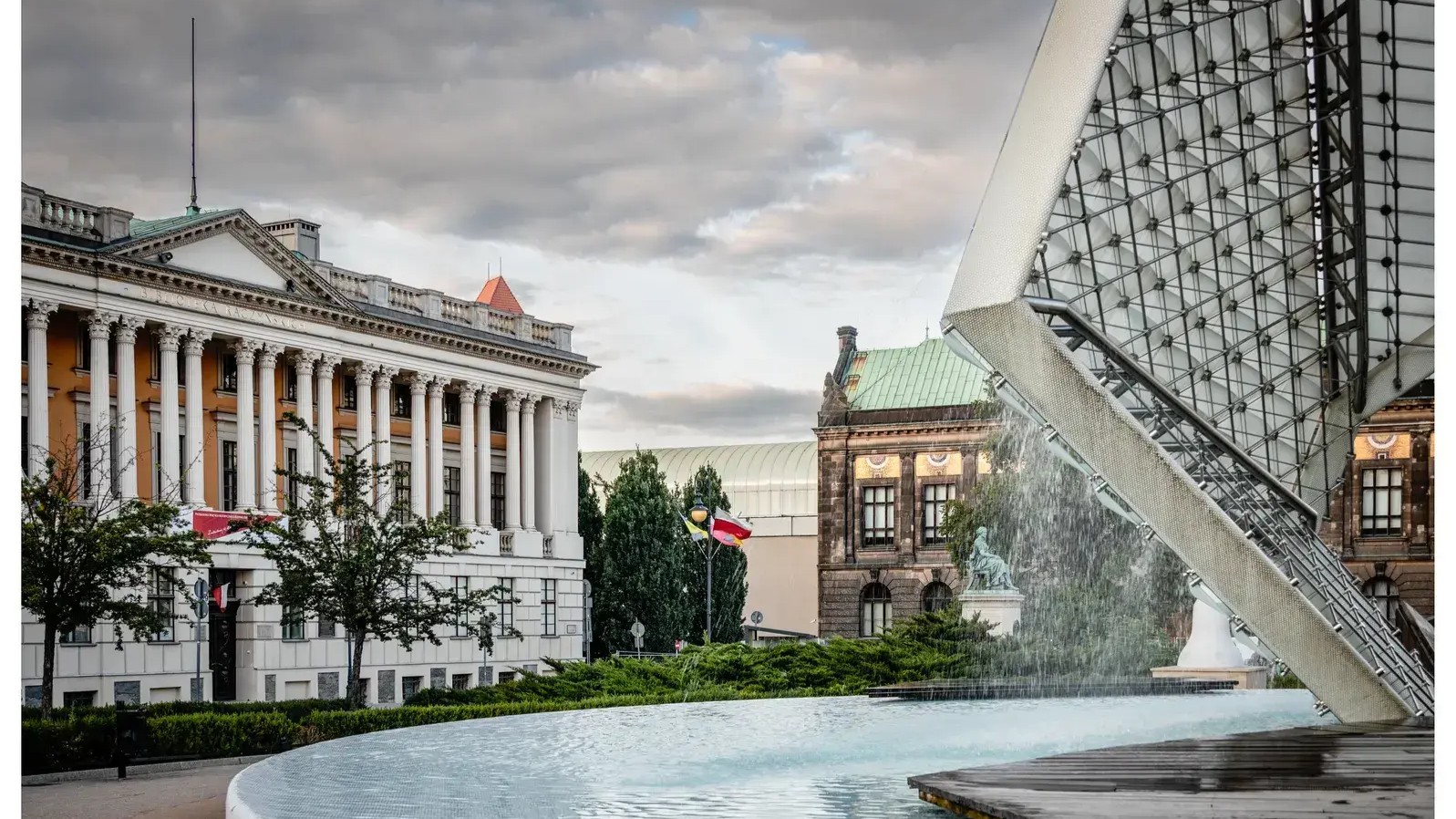 Biblioteka Raczyńskich przy pl. Wolności w Poznaniu. Widoczna na tle placu z fontanną i charakterystyczną konstrukcją zadaszenia. W tle znajdują się zabytkowe budynki oraz pomnik. Nad miastem zachmurzone niebo.