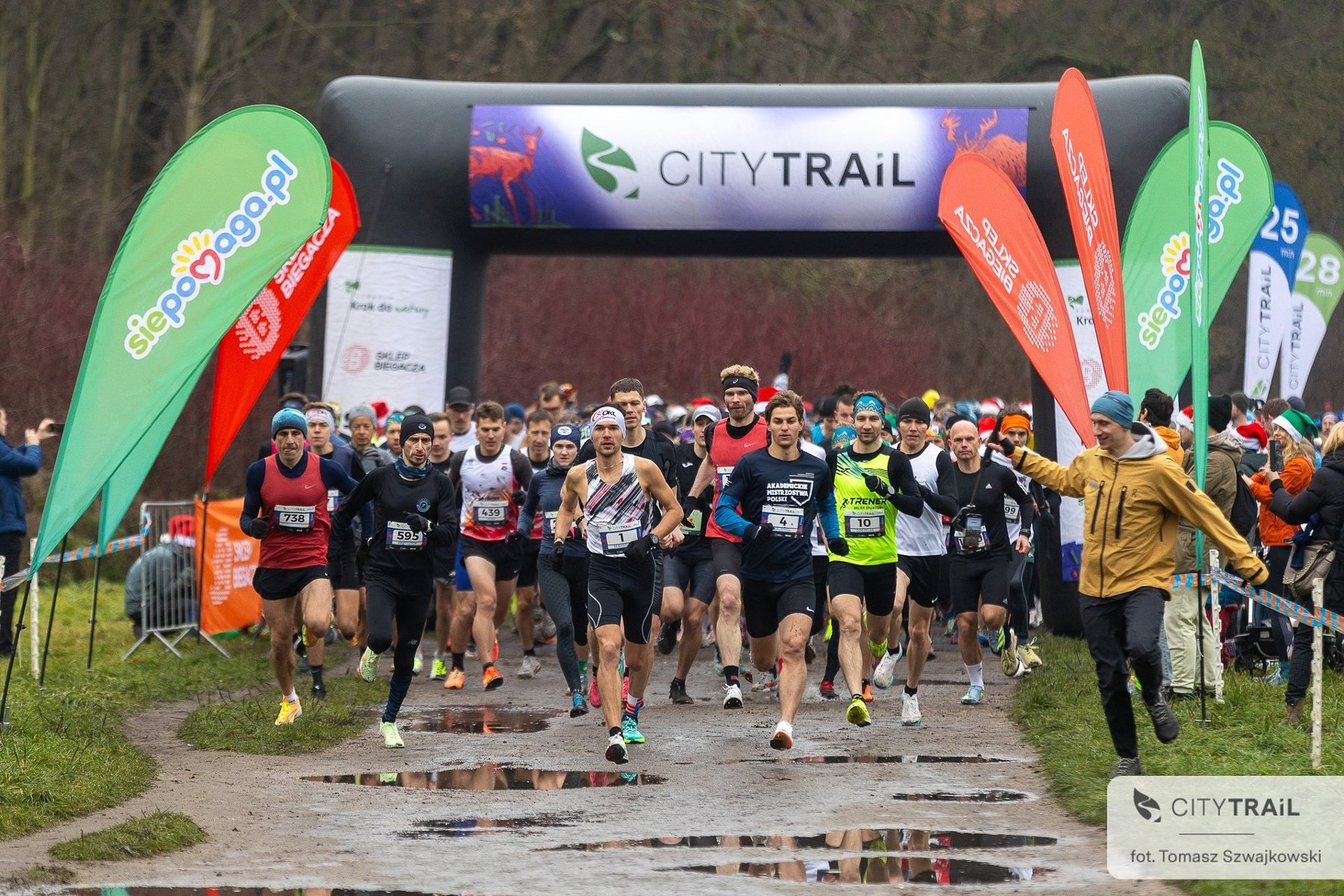 Bieg City Trail w Poznaniu, fot. Tomasz Szwajkowski - duża grupa biegaczy rozpoczyna bieg. W tle widać las i bramę startową oraz inne nośniki