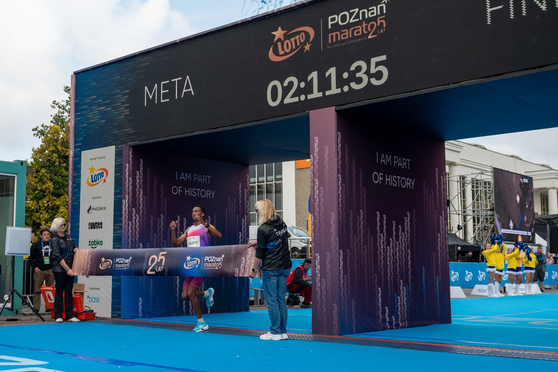 Biegacz przecinający wstęgę mety podczas 25. Poznań Maratonu pod czarną bramą mety z cyfrowym zegarem pokazującym czas biegowy.