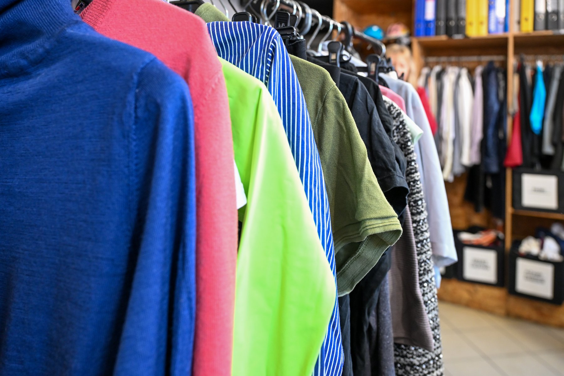 Bliskie ujęcie rzędu kolorowych ubrań zawieszonych na czarnych wieszakach wewnątrz sklepu lub magazynu. Na pierwszym planie widoczny jest niebieski sweter z golfem, obok którego wiszą ubrania w kolorach czerwonym, neonowym zielonym, niebieskim w białe paski oraz oliwkowym. Tło jest lekko rozmyte, ukazując dalsze regały z odzieżą oraz segregatory na półkach, co sugeruje zaplecze organizacyjne lub sklep z odzieżą używaną.