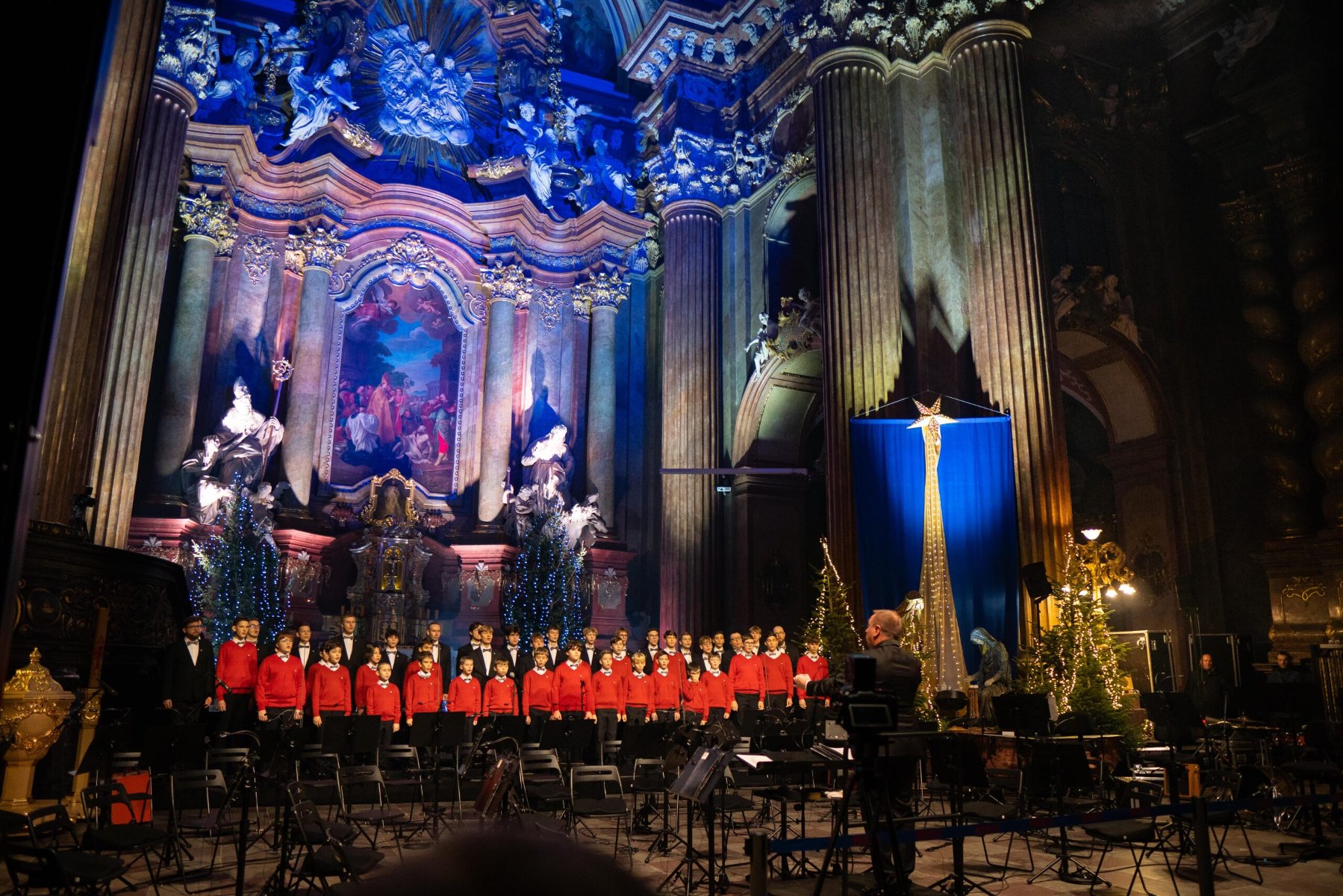 Bogato zdobiony ołtarz w kościele. Pod nim stoi chór męski. W pierwszym rzędzie stoją chłopcy w czerwonych swetrach, białych koszulach i czarnych spodniach. Za nim stoją chlopcy w czarnych garniturach. Przed chłopcami stoi dyrygent ubrany na czarno. Ołtarz jest rozświetlony żółtym i niebieskim światłem.