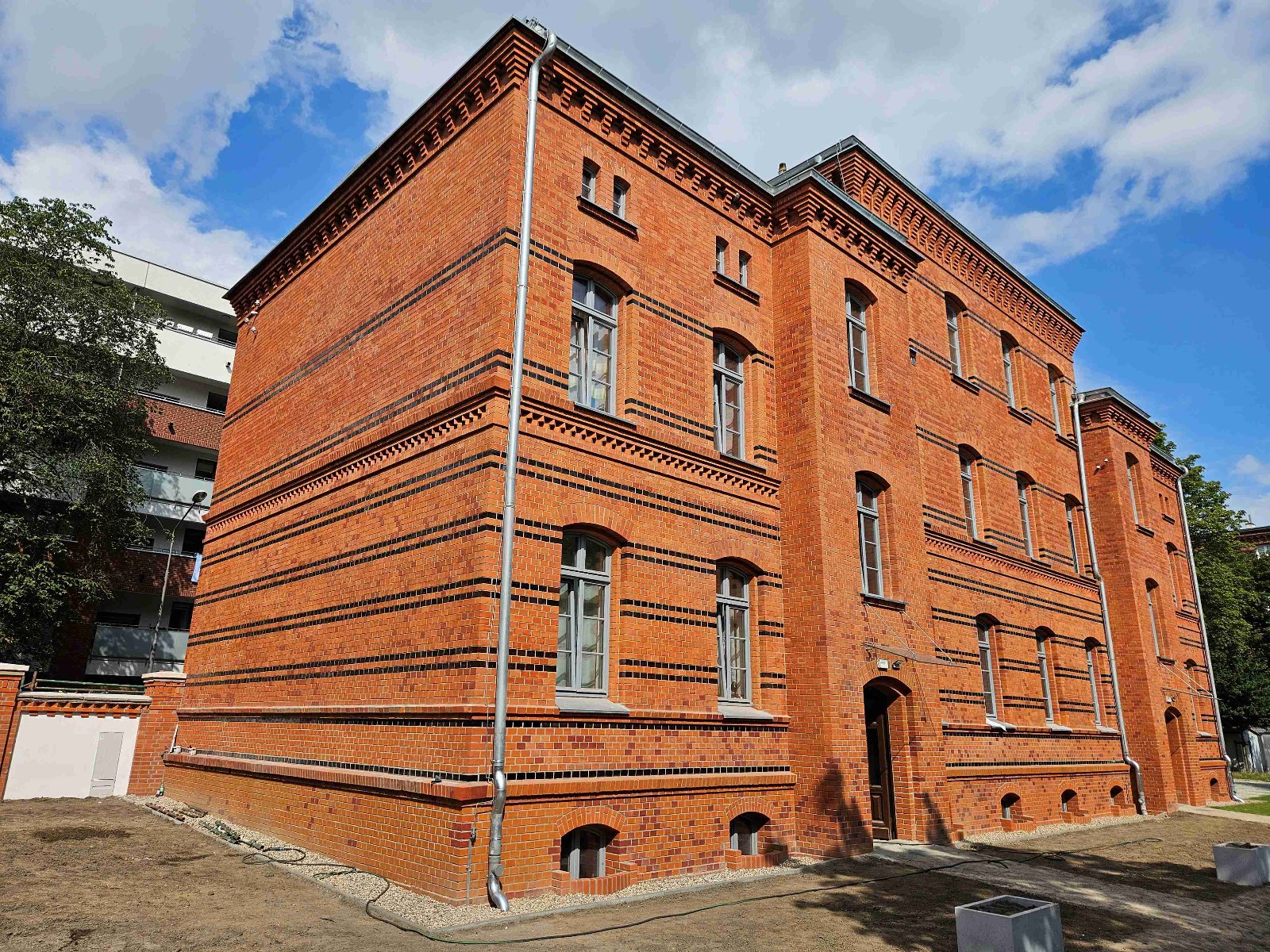 Budynek z czerwonej cegły, o prostokątnym kształcie i oknami z szarymi ramami. Fasada jest zdobiona czarnymi paskami z cegły. Przed budynkiem widoczny jest trawnik.