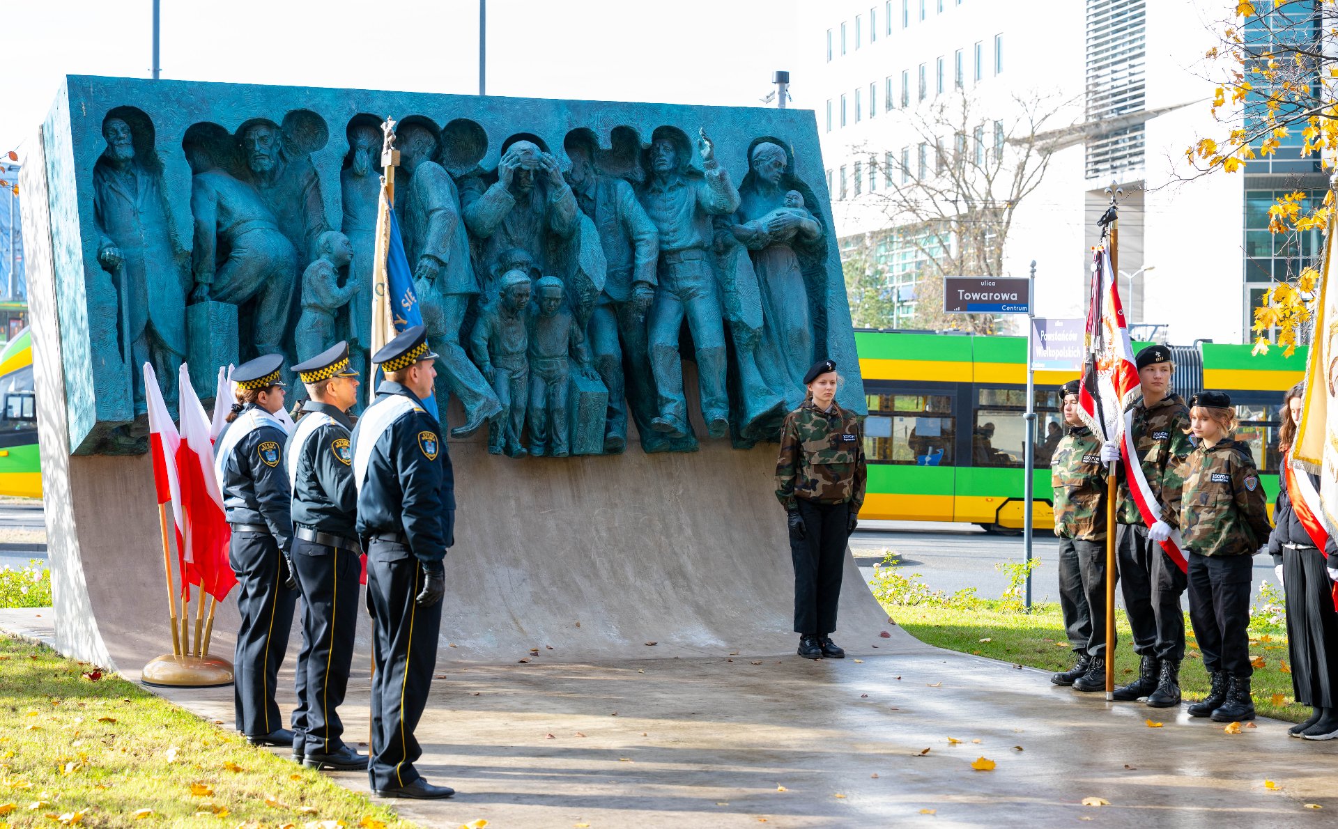 Ceremonia przed pomnikiem: Grupa umundurowanych funkcjonariuszy stoi na baczność po lewej stronie, z flagami. Po prawej stronie, również na baczność, stoi grupa młodzieży w mundurach maskujących, trzymająca sztandary. W tle, na ulicy, przejeżdża duży, zielony tramwaj.