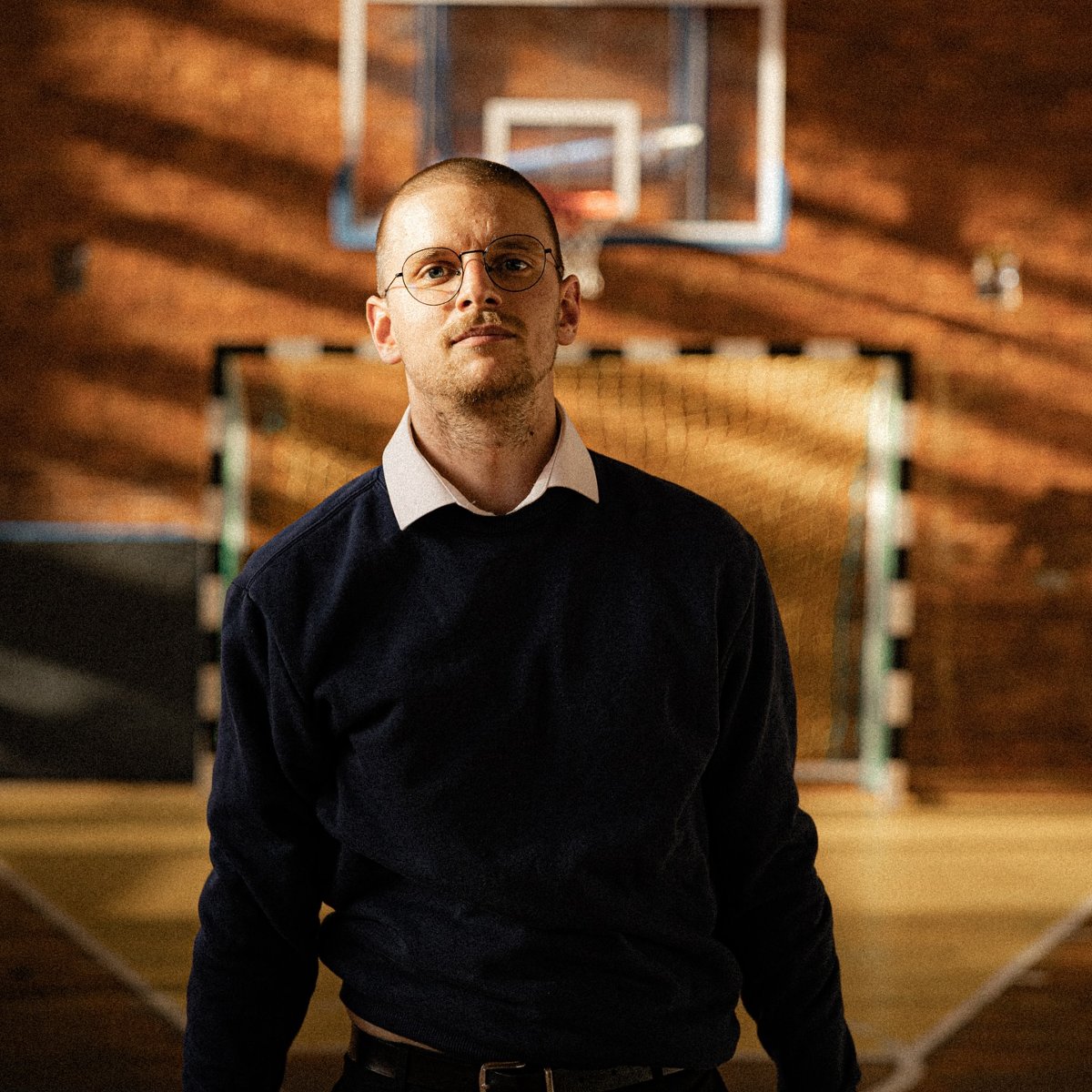 Chłopak w swetrze i okularach stoi na sali sportowej.