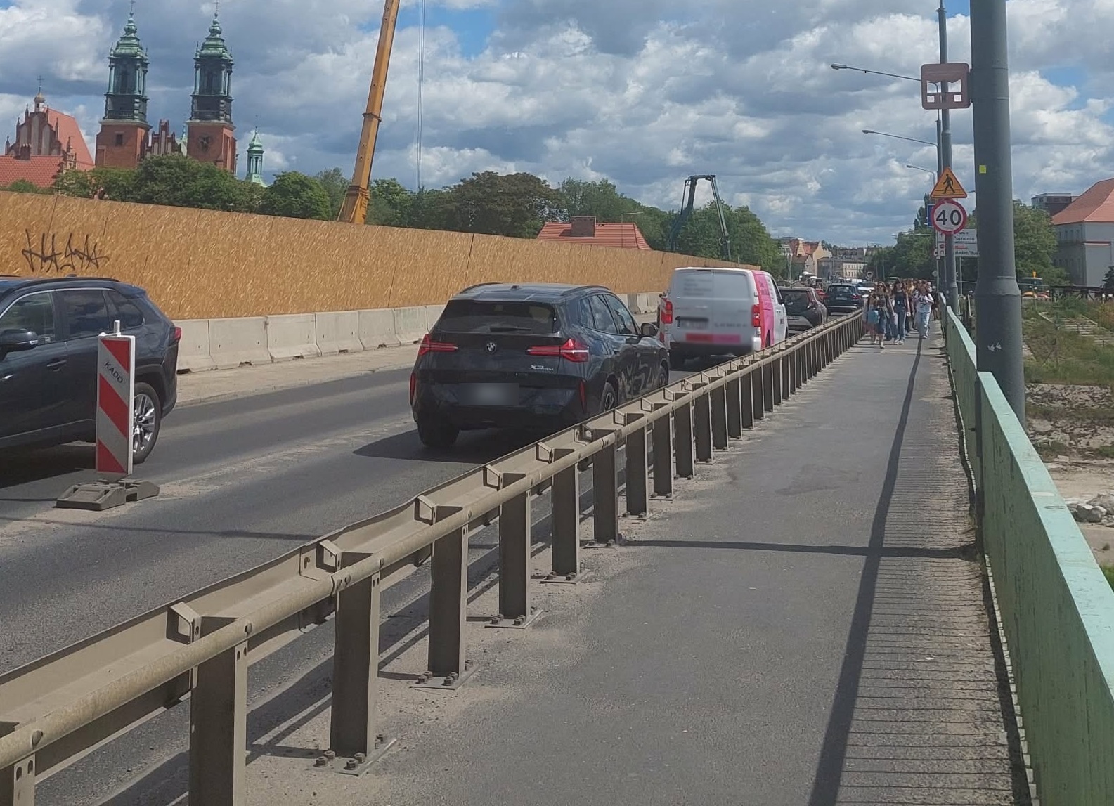Chodnik przy jezdni, obok przejeżdżające samochody. W tle budynek kościoła. Widoczne maszyny budowlane.