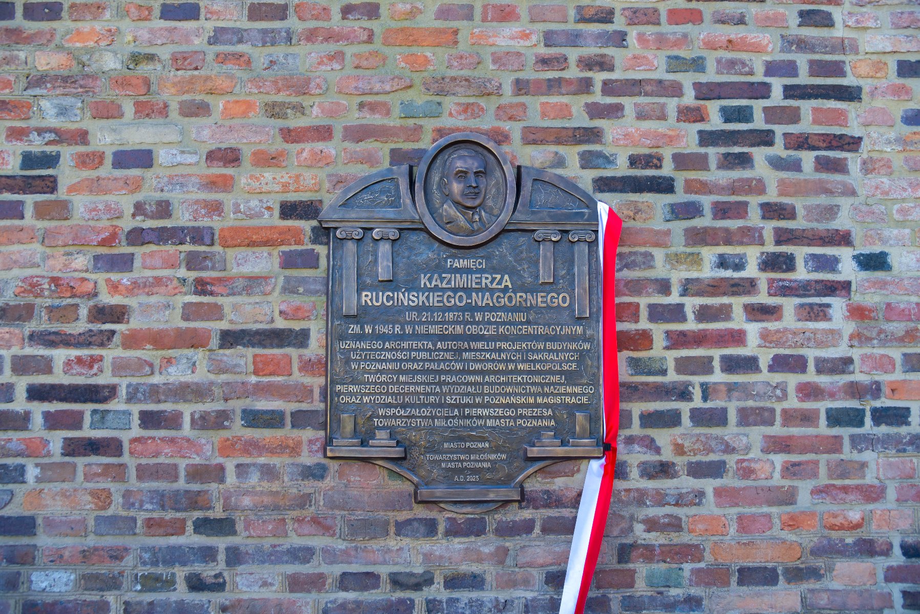 Ciemna, metalowa tablica pamiątkowa zamontowana na ceglanym murze. U góry tablicy znajduje się płaskorzeźba z portretem mężczyzny. Poniżej widnieje napis: "PAMIĘCI KAZIMIERZA RUCIŃSKIEGO-NAGÓRNEGO", a pod nim szczegółowy opis jego zasług jako architekta. Z prawej strony tablicy przewieszona jest biało-czerwona wstęga.