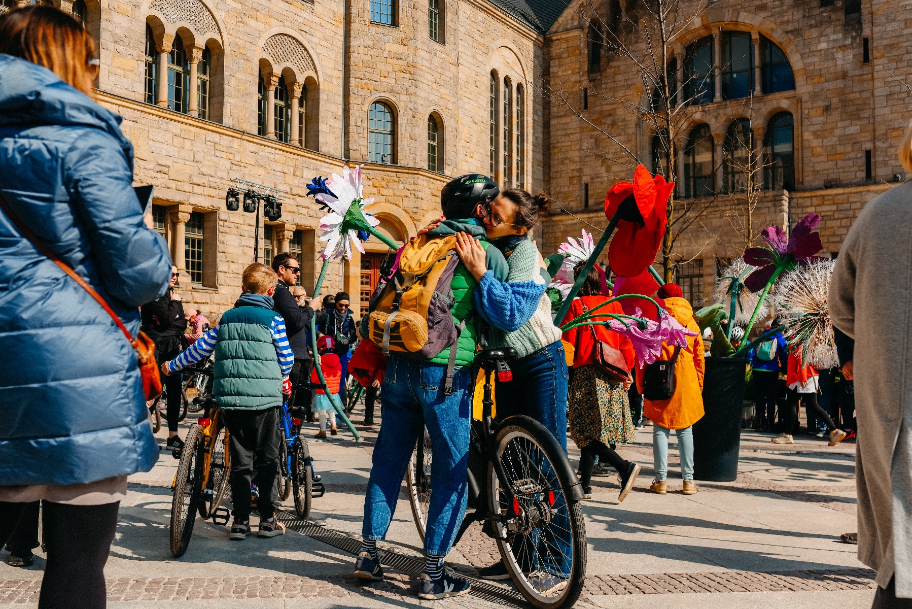 Ciepła scena na placu przed monumentalnym, kamiennym gmachem Centrum Kultury Zamek w Poznaniu. Na środku uwieczniono czuły uścisk dwojga rowerzystów: kobiety w niebieskim swetrze i mężczyzny w kasku z żółtym plecakiem. Wokół nich znajdują się ogromne, kolorowe sztuczne kwiaty - maki i stokrotki - oraz grupa ludzi z rowerami, w tym dziecko w kamizelce. Zdjęcie wykonane w słoneczny dzień, emanuje radosną, wiosenną atmosferą.