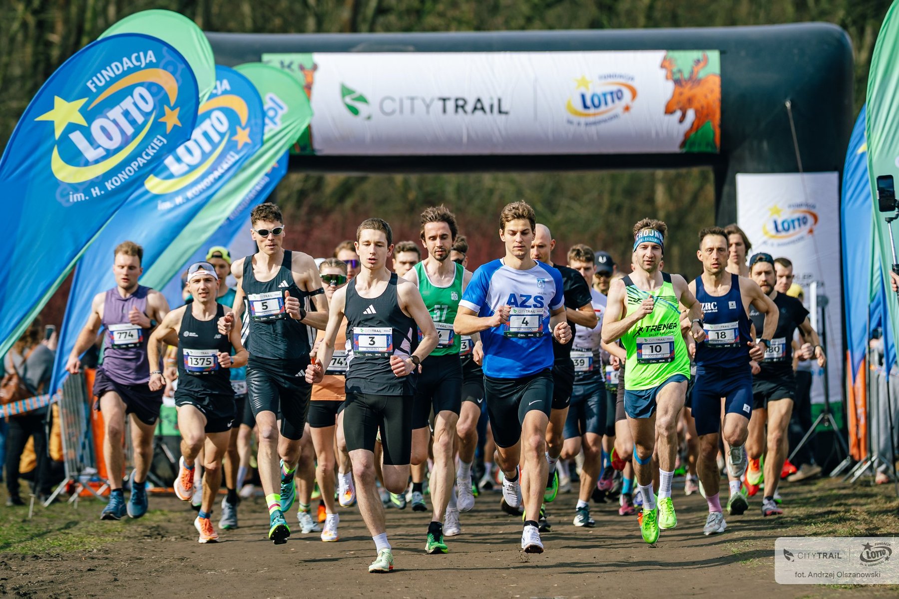 CITY TRAIL w Poznaniu, fot. Andrzej Olszanowski - kilkudziesięciu biegaczy w sportowych strojach biegnie po starcie. Za nimi jest brama pneumatyczna i las