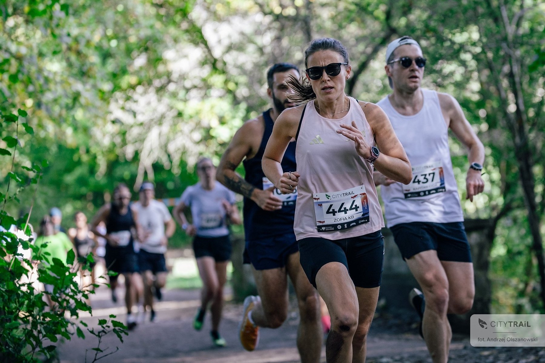 Cykl City Trail w Poznaniu, fot. Andrzej Olszanowski - na pierwszym planie trójka biegaczy w krótkich spodniach i koszulkach (1 kobieta oraz 2 mężczyzn) w tle kilkoro kolejnych biegaczy, a także drzewa z zielonymi liśćmi