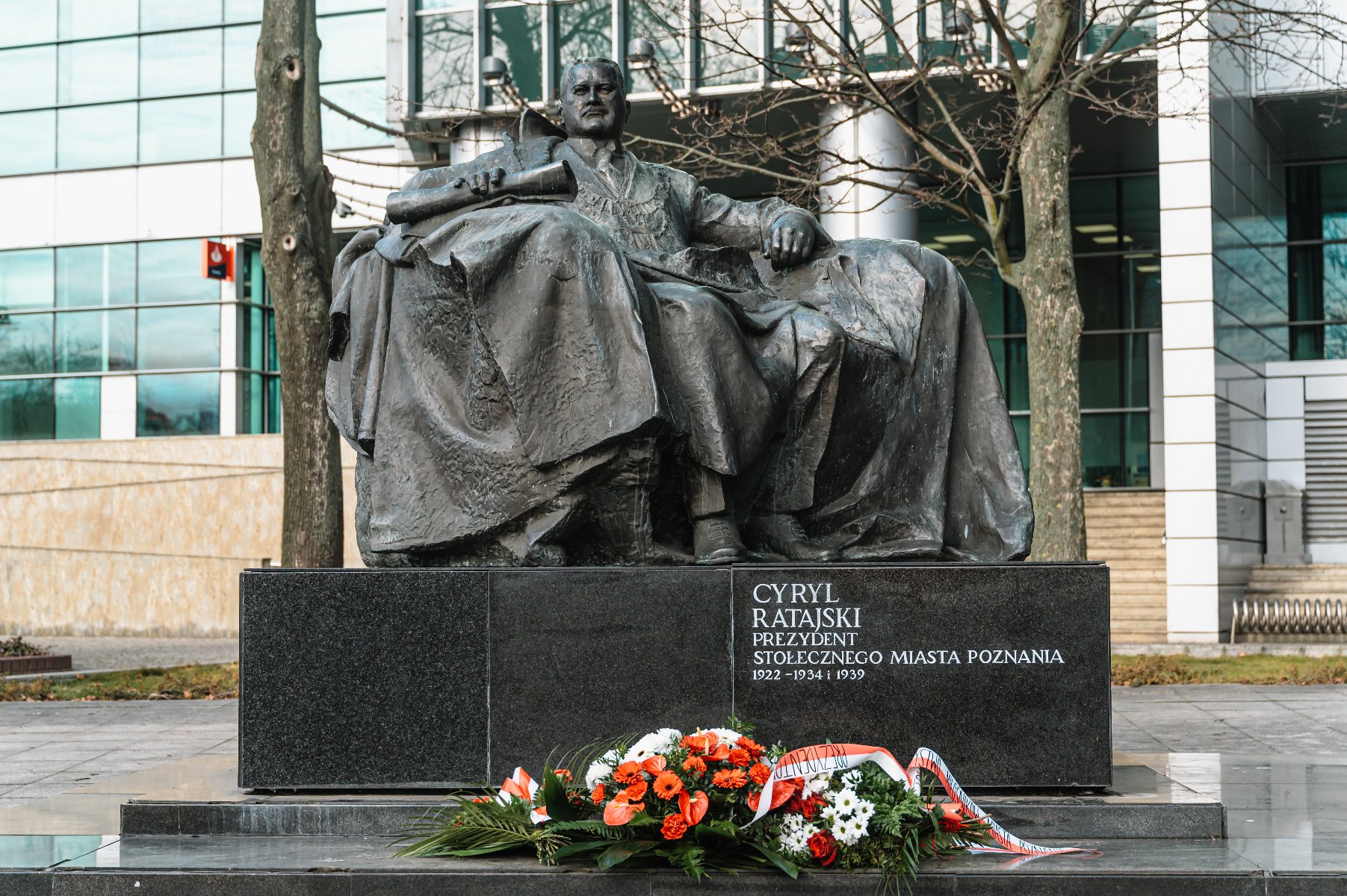 Cyryl Ratajski siedzący na pomniku z brązu na czarnym cokole. Przed cokołem ułożone są kwiaty w kolorach białym, czerwonym i zielonym oraz wstążka. Na cokole widnieje napis: "CYRYL RATAJSKI PREZYDENT STOŁECZNEGO MIASTA POZNANIA 1922 - 1934 I 1939".