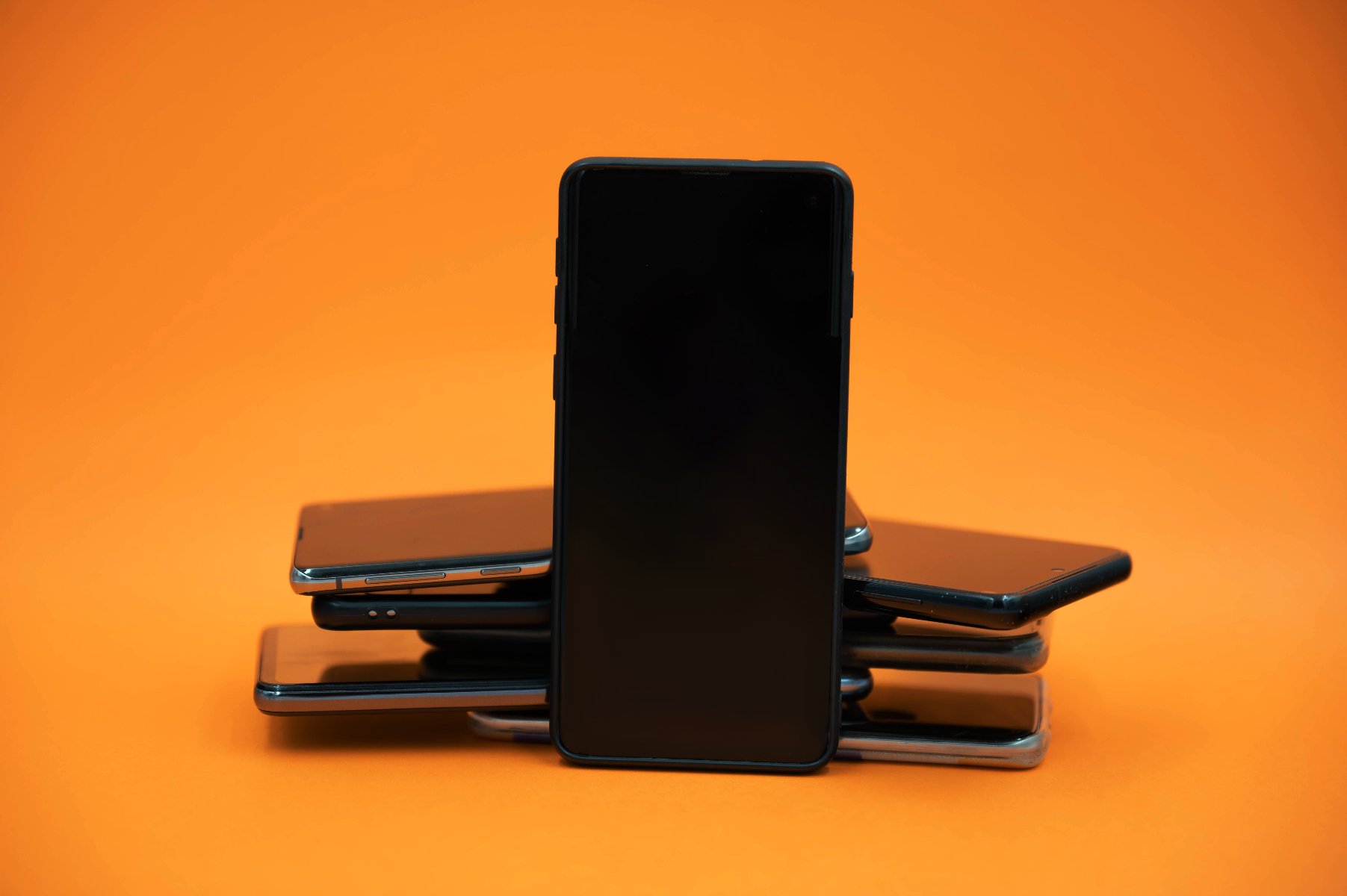 Czarny smartfon stojący pionowo na tle ułożonych poziomo innych smartfonów. Wszystko na pomarańczowym tle.