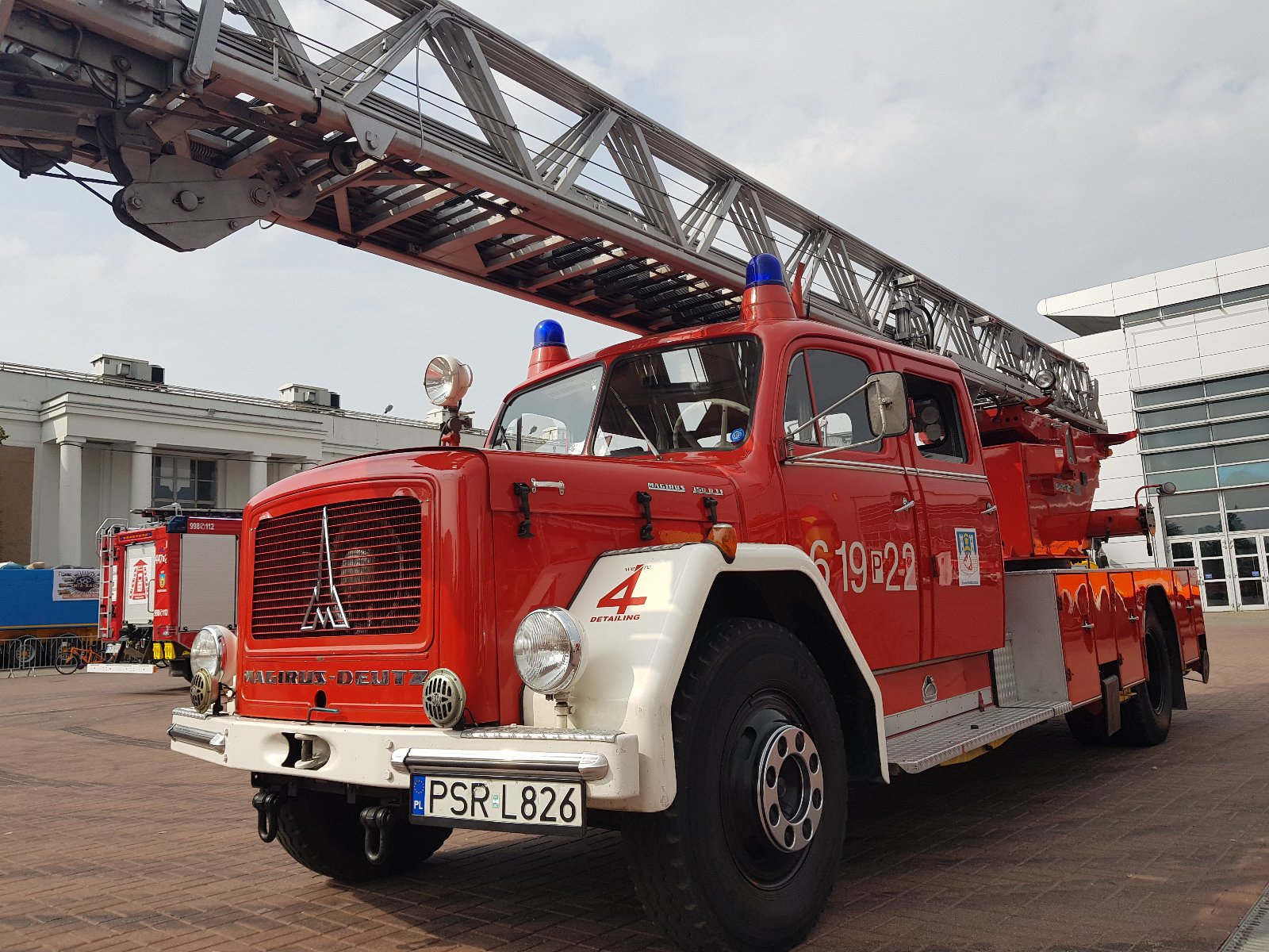 Czerwony zabytkowy wóz strażacki marki Magirus-Deutz z drabiną mechaniczną i numerem taktycznym 619[P]22 zaparkowany na placu przed nowoczesnym budynkiem.