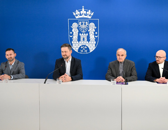 Czterech mężczyzn siedzących za białym stołem prezydialnym podczas konferencji prasowej, na tle niebieskiej ścianki z herbem Miasta Poznania.