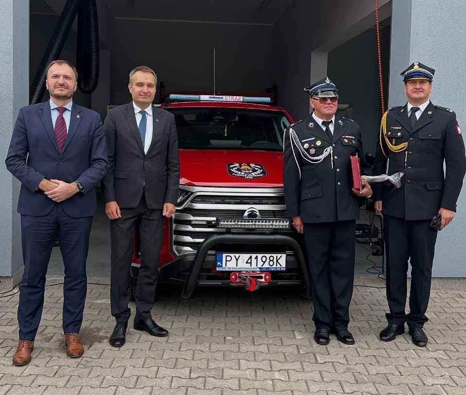 czterwch mężczyzn dwóch w garnirurach i dwóch w galowych mundurach strażaków pozuje do zdjęcia na tle terenowego samochodu strażackiego