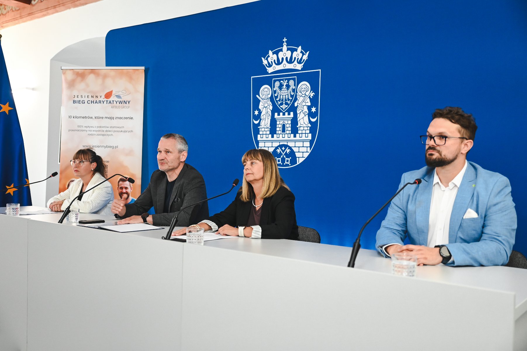 Cztery osoby siedzą przy stole konferencyjnym podczas oficjalnej konferencji prasowej. Za nimi znajduje się niebieska ścianka z herbem miasta Poznania oraz flaga Unii Europejskiej. Po lewej stronie stoi roll-up promujący Jesienny Bieg Charytatywny. Osoby mają przed sobą mikrofony i szklanki z wodą, wyglądają na skupione i zaangażowane w omawiane tematy