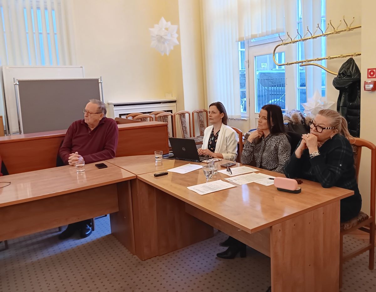 cztery osoby siedzą w sali przy stole konferencyjnym, patrzą w jedną stronę