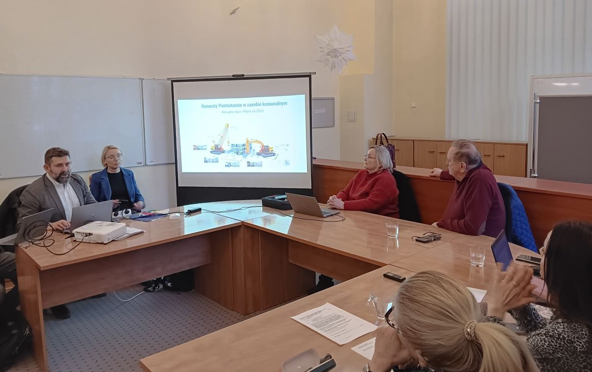 cztery osoby siedzą w sali przy stole konferencyjnym, patrzą w jedną stronę