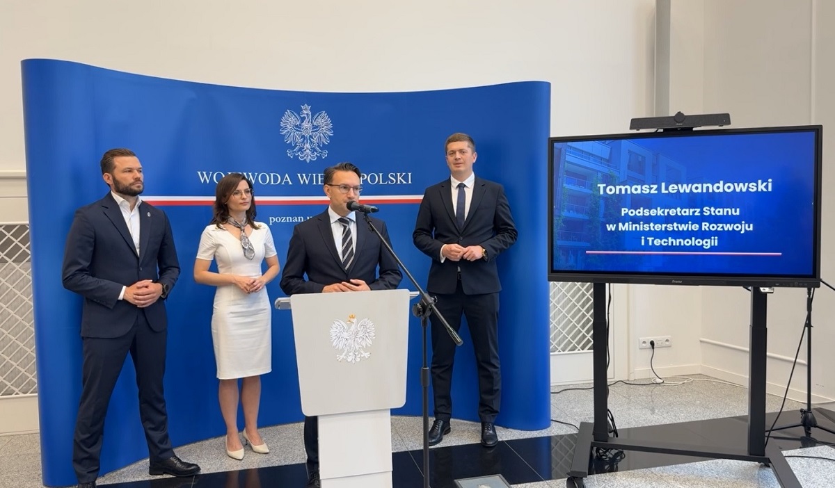 cztery osoby stoją na konferencji prasowej, trzech mężczyzn o kobieta, mężczyźni ubrani w ciemne garnitury, kobieta w białej sukni, jeden z mężczyzn stoi przy mównicy z mikrofonem