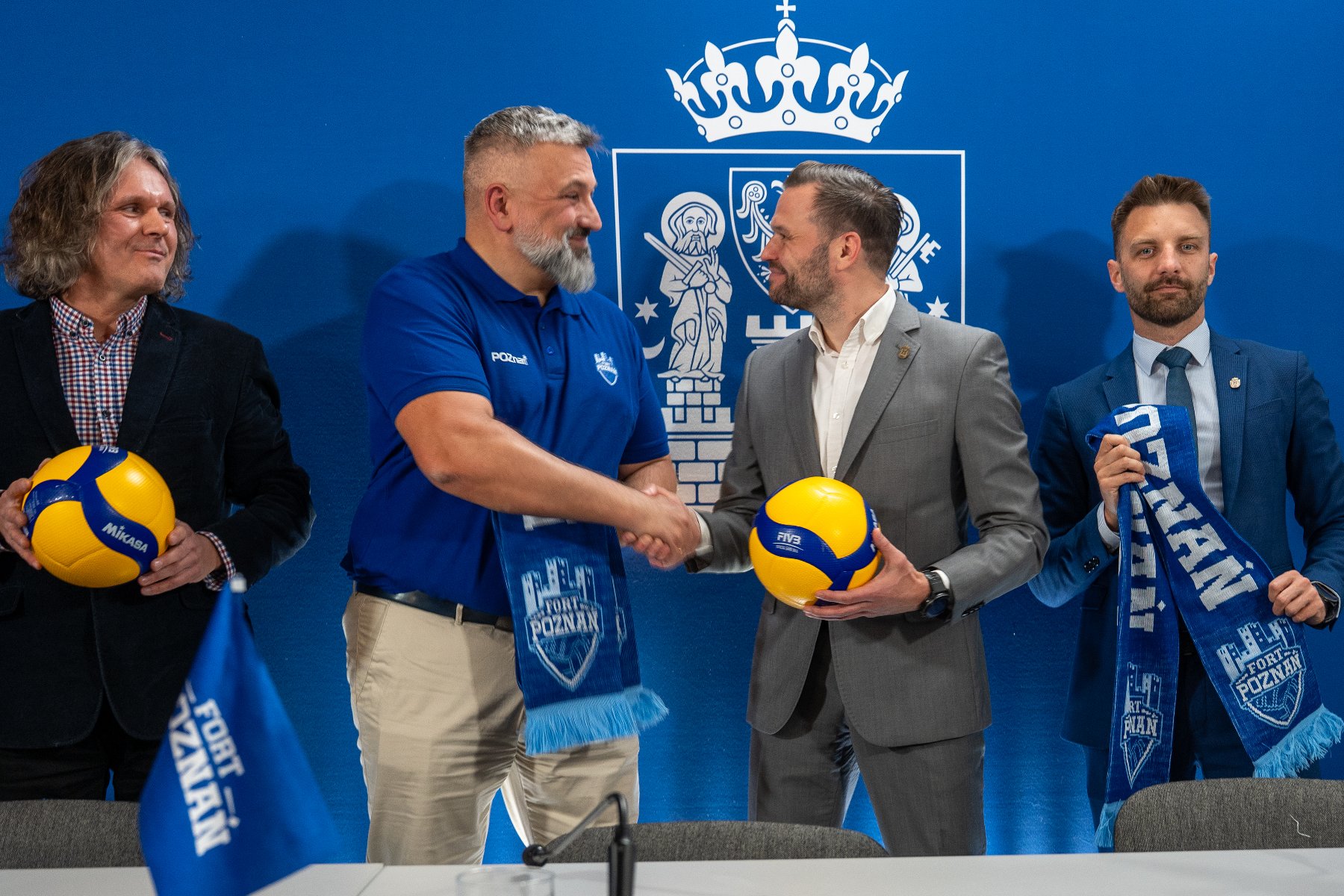 Cztery osoby stoją po konferencji trzymając piłki siatkowe i szaliki sportowe. Dwóch mężczyzn na środku podaje sobie ręce