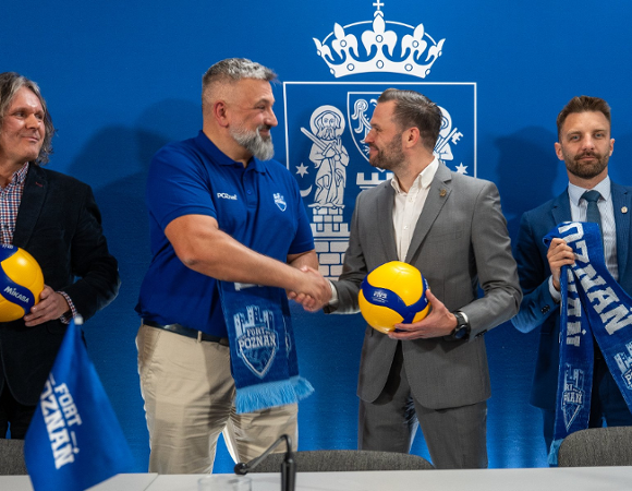 Cztery osoby stoją po konferencji trzymając piłki siatkowe i szaliki sportowe. Dwóch mężczyzn na środku podaje sobie ręce