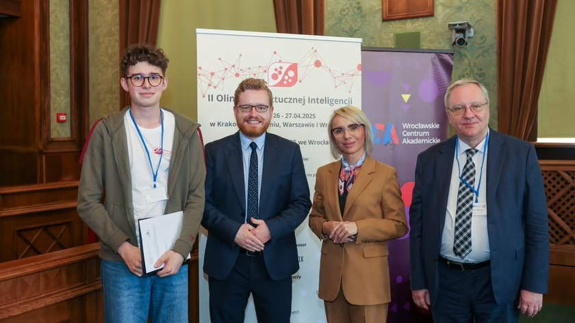 Cztery osoby stojące obok siebie na tle banera informującego o II Olimpiadzie Sztucznej Inteligencji. Od lewej: młody mężczyzna w okularach i jasnej bluzie z identyfikatorem na szyi, uśmiechnięty mężczyzna w garniturze z krawatem, kobieta w brązowym garniturze z chustą na szyi i okularach oraz starszy mężczyzna w garniturze z krawatem i okularach. W tle po prawej widoczne jest logo Wrocławskiego Centrum Akademickiego.