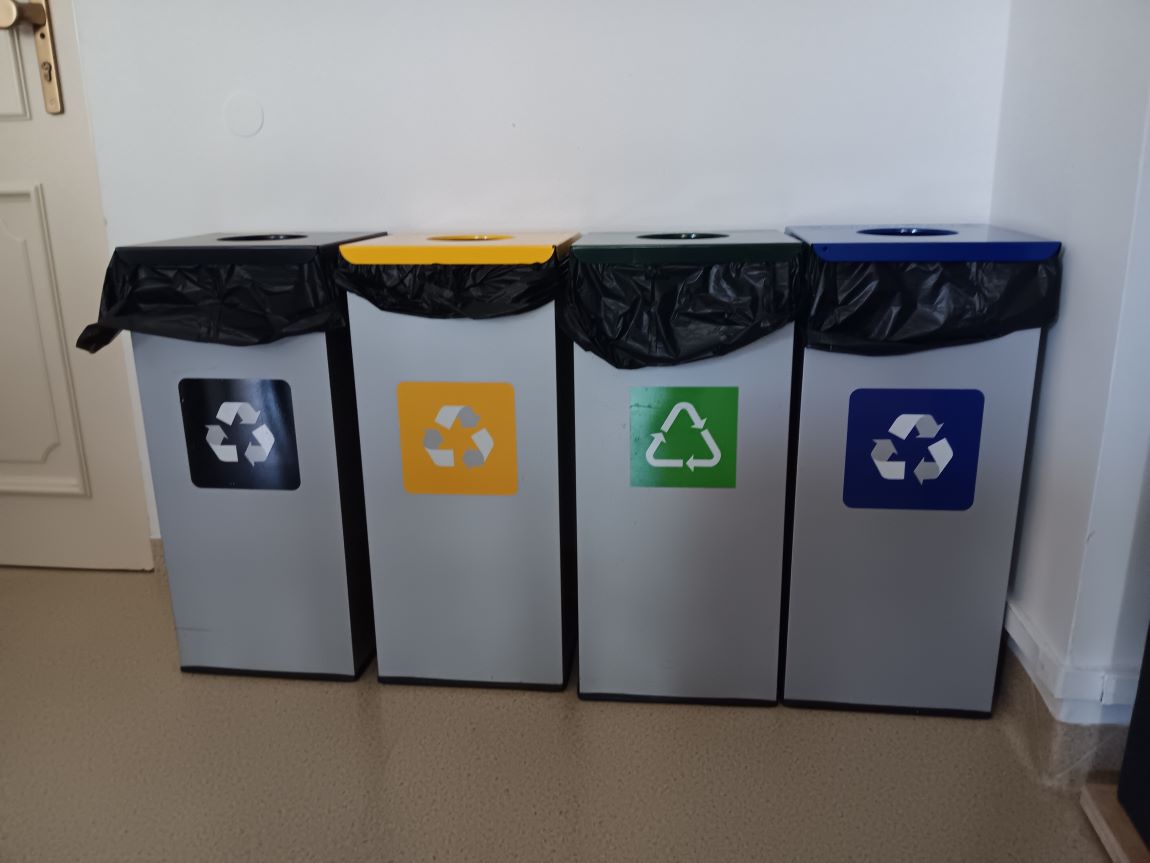 cztery pojemniki do segregacji różnego rodzaju odpadów, stoją w rzędzie przy ścianie