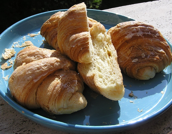 Cztery złociste, świeże rogaliki na turkusowym talerzu. Jeden z rogalików jest przekrojony na pół, ukazując jego jasny, puszysty środek. Wokół rogalików widać okruszki. Talerz stoi na kamiennym blacie w ostrym słońcu.