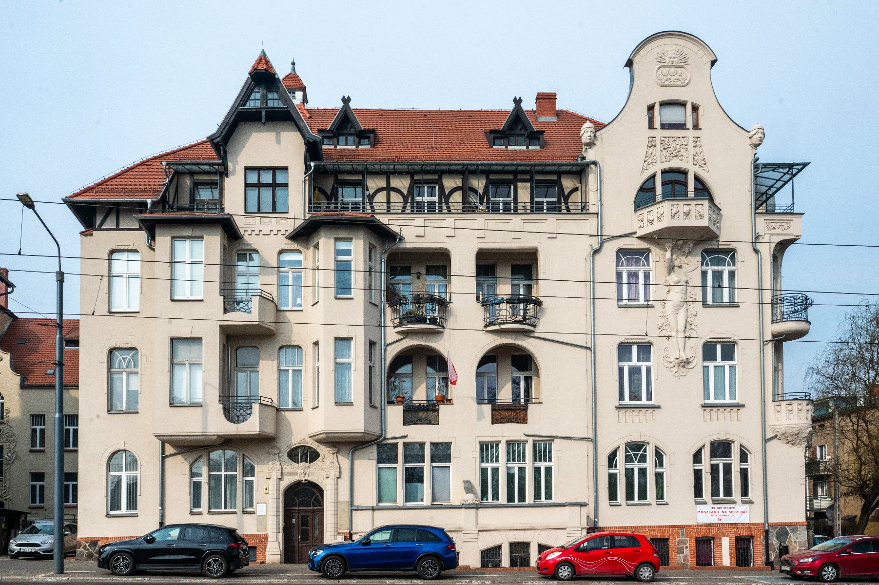 Detal architektoniczny nad drzwiami. Konstrukcja zawiera misternie rzeźbione wzory roślinne i liściaste w kamieniu lub tynku. Nad drzwiami znajduje się łukowe okno podzielone na trzy sekcje pionowymi prętami. Elementy dekoracyjne obejmują symetryczne wzory roślinne po obu stronach łuku oraz centralny motyw roślinny na szczycie łuku. Po lewej stronie drzwi widoczna jest mała tabliczka z numerem "4".