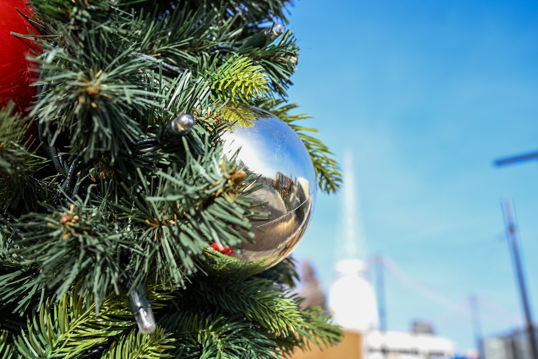 Detal choinki ozdobionej lampkami i dużą, lśniącą srebrną bombką, w której odbija się otoczenie. W tle widoczne jest intensywnie niebieskie niebo w słoneczny dzień.