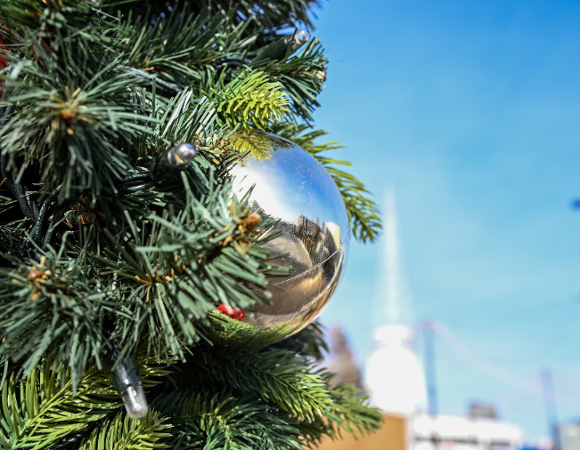 Detal choinki ozdobionej lampkami i dużą, lśniącą srebrną bombką, w której odbija się otoczenie. W tle widoczne jest intensywnie niebieskie niebo w słoneczny dzień.