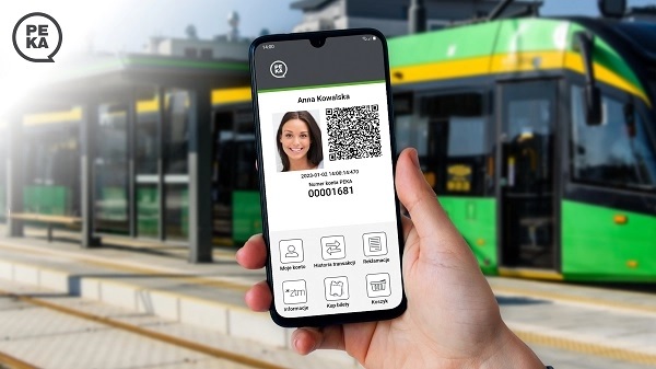 dłoń trzyna telefon komórkowy, na ekranie wyświetona twarz kobiety i QR kod, w tle za telefonem wiata przystankowa i tramwaj