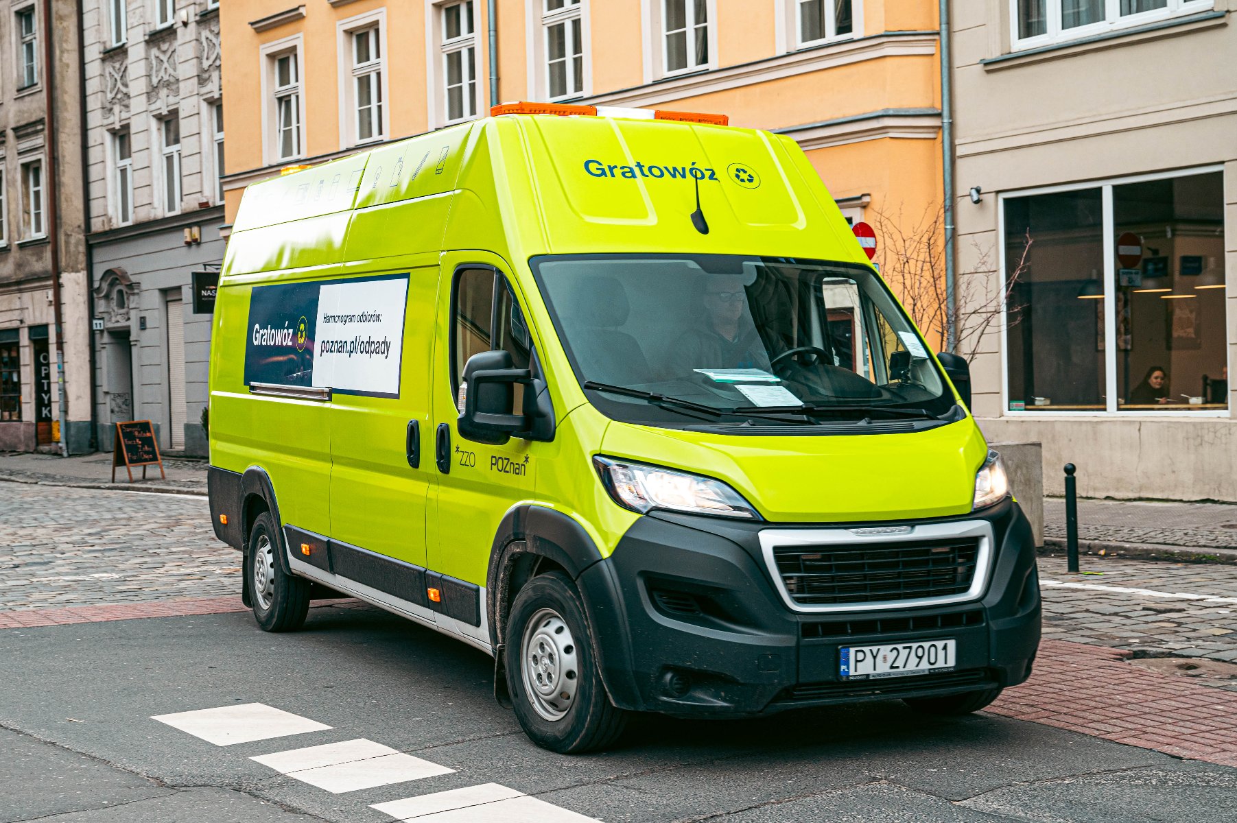Dostawczy samochód marki Peugeot w jaskrawożółtym, seledynowym kolorze, jadący po brukowanej ulicy w mieście. Na dachu samochodu pomarańczowe lampy i duży, zielony napis "Gratowóz". Po bokach reklama i adres strony, w tym: "poznan.pl/odpady".