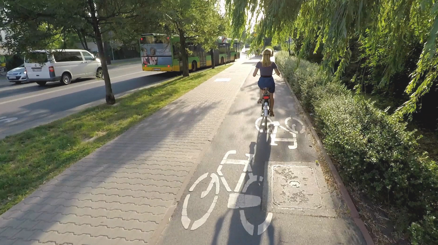 droga rowerowa i chodnik, na drodze rowerowej jedzie rowerzystka, po obu stronach zieleń - drzewa i krzewy, na ulicy jadą dwa samochody i autobus komunikacji miejskiej