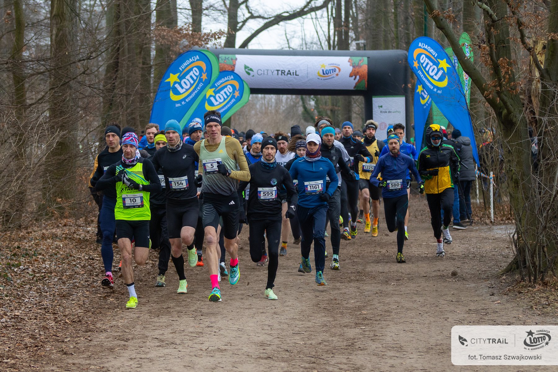 Duża grupa biegaczy w strojach sportowych startuje w biegu przełajowym na leśnej ścieżce. Zawodnicy na pierwszym planie mają przypięte numery startowe, a wielu z nich nosi czapki i opaski, co sugeruje chłodną pogodę. W tle widoczna jest dmuchana brama startowa z napisem "CITY TRAIL" oraz niebieskie flagi reklamowe z logo Fundacji Lotto. Zdjęcie wykonane w lesie w bezlistnym okresie roku.