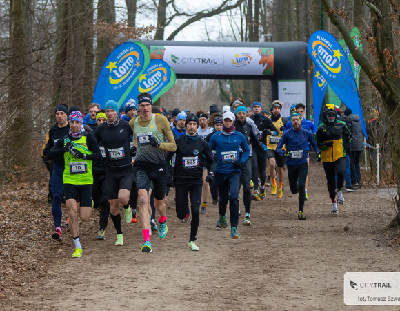 Duża grupa biegaczy w strojach sportowych startuje w biegu przełajowym na leśnej ścieżce. Zawodnicy na pierwszym planie mają przypięte numery startowe, a wielu z nich nosi czapki i opaski, co sugeruje chłodną pogodę. W tle widoczna jest dmuchana brama startowa z napisem "CITY TRAIL" oraz niebieskie flagi reklamowe z logo Fundacji Lotto. Zdjęcie wykonane w lesie w bezlistnym okresie roku.