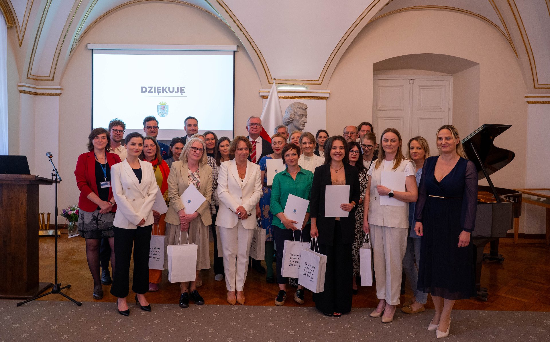 Duża grupa ludzi, głównie kobiet w eleganckich strojach, pozuje do zdjęcia. W tle widoczny jest ekran z napisem "DZIĘKUJĘ", a także popiersie na postumencie i fragment fortepianu. Niektórzy z uczestników trzymają w rękach białe torby i certyfikaty.