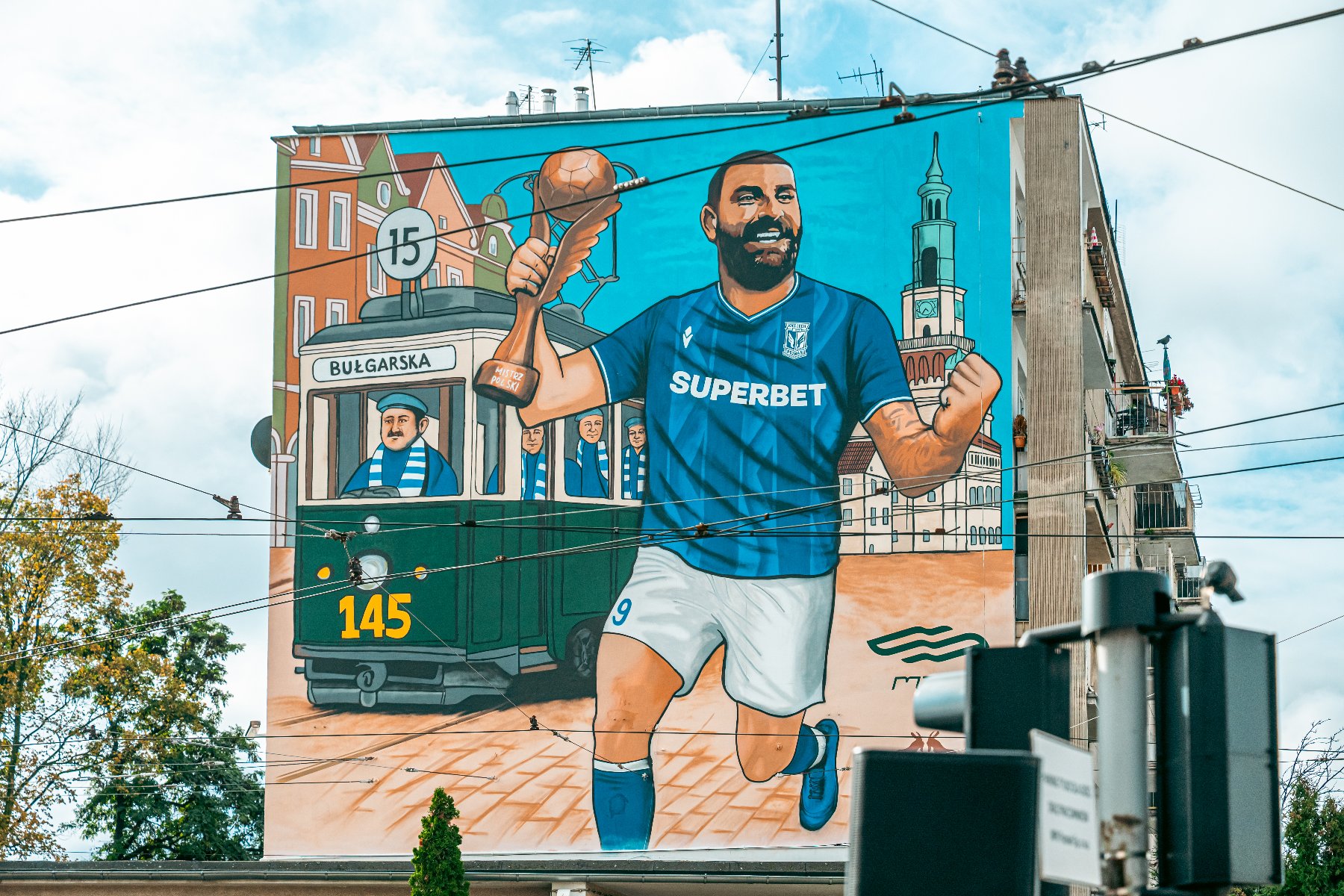 Duży, kolorowy mural na bocznej ścianie budynku, namalowany w stylu komiksowym, przedstawiający sportowca i scenę z miasta. W centrum widać uśmiechniętego piłkarza w niebieskiej koszulce z napisem "SUPERBET" i numerem 9, który biegnie, trzymając w górze złotą statuetkę w kształcie piłki nożnej. W tle widoczny jest zielony tramwaj z numerem 145 i napisem "BULGARSKA", prowadzony przez motorniczego, oraz zabudowania miejskie (kamienice i Wieża Ratusza).