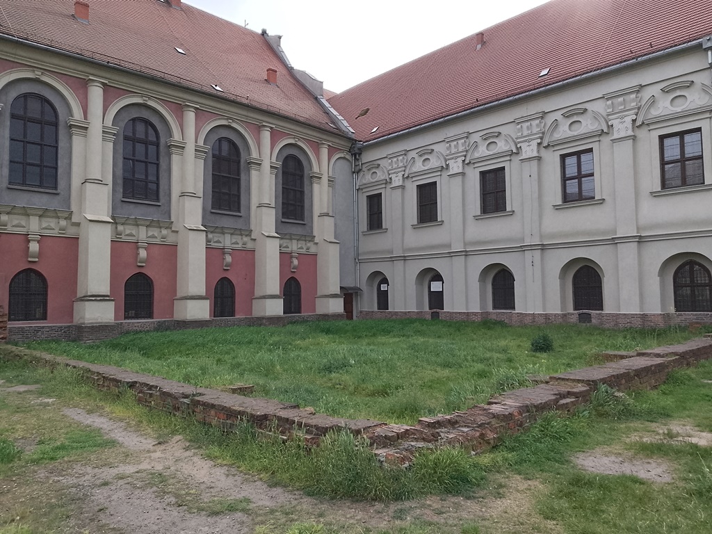 dwa zabytkowe budynki ze spadzistymi dachami, wysokimi oknami zdobionymi łukami, przed nimi teren porośnięty trawą i pozostałości murów lub ścian budynków